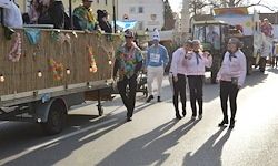 Eine Gruppe von Menschen in Kostümen geht die Straße entlang, im Hintergrund befinden sich dekorierte Fahrzeuge.