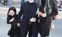 Eine Familie in Halloween-Kostümen steht auf der Straße. Ein Kind in einem Hexenkostüm und zwei Erwachsene in schwarzen Outfits. Der Erwachsene in der Mitte hält ein Ei.