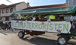 Ein Anhänger mit einem grünen Baldachin steht auf der Straße. Ein Banner mit dem Wort 'Klimaterrorristen' ist am Anhänger angebracht. Menschen sitzen im Inneren des Anhängers.