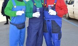 Drei Männer in Mario-Kostümen stehen auf einem Parkplatz, jeder mit einem Becher in der Hand. Einer trägt Grün, der andere Rot und der Dritte Blau. Ein Van ist im Hintergrund zu sehen.