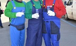 Drei Personen in Mario- und Luigi-Kostümen stehen draußen. Jede hält einen Becher. Ein Van ist im Hintergrund geparkt.