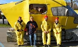Fünf Personen in gelben Schutzanzügen stehen vor einem gelben Objekt, wahrscheinlich einem U-Boot. Ein Mann in einem schwarzen Hemd steht zwischen zwei Männern mit roten Mützen.