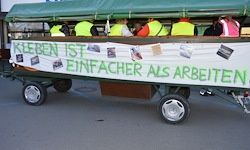 Ein Anhänger mit einem grünen Dach transportiert Menschen in neonfarbenen Westen. Ein Banner lautet 'Kreben ist einfacher als arbeiten.'