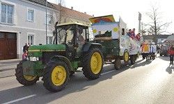 Ein grüner Traktor mit gelben Rädern und einem dekorierten Anhänger fährt auf einer Straße, wobei Menschen auf der Seite gehen. Im Hintergrund sind Gebäude und Bäume zu sehen.