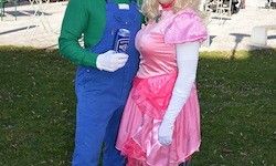 Zwei Personen, die als Mario und Prinzessin Peach gekleidet sind, stehen auf einem Rasen mit grünem Gras und einem verschwommenen Hintergrund.
