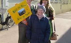 Drei Personen stehen auf der Straße. Die mittlere Person hält ein gelbes Schild mit Text. Die Person auf der linken Seite hält dasselbe Schild. Die Person auf der rechten Seite trägt ein grünes Kleid.