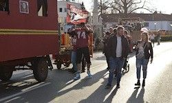 Eine Gruppe von Menschen geht auf einer Straße mit einem roten Lkw und einem Wagen in der Nähe. Ein Mann in einem roten Trikot hält eine Flagge. Ein Kinderwagen wird geschoben, und andere gehen dahinter.