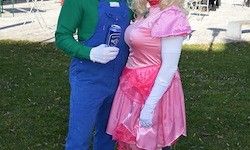 Eine Person in einem Mario-Kostüm und eine weitere in einem rosa Prinzessinnen-Kostüm stehen auf Gras. Der Mario-Charakter hält eine Dose.