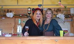 Zwei Frauen in Kostümen stehen hinter einer Holzbar. Eine hält ein Weinglas und die andere einen grünen Becher. Verschiedene Gegenstände, darunter Flaschen und ein Kronleuchter, befinden sich auf der Bar und den Wänden.