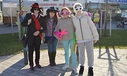 Vier Personen stehen auf einem gepflasterten Bürgersteig. Sie tragen verschiedene Kostüme und Accessoires wie Hüte, Brillen und Handschuhe. Im Hintergrund befinden sich ein Gebäude, einige geparkte Fahrzeuge und ein Rasenbereich.