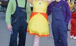 Drei Menschen in Kostümen stehen draußen zusammen. Ein Mann in grüner Arbeitskleidung, eine Frau in einem gelben Kleid und ein Mann in blauer Arbeitskleidung.