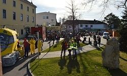 Eine Gruppe von Menschen in gelben Anzügen geht auf einem Bürgersteig. Ein gelber Bus und mehrere Gebäude sind im Hintergrund zu sehen.
