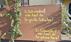 Ein braunes Schild mit gelbem Text lautet 'Ei. Schwadorf, was hast du fur groBe Schutze!' und 'Ei. Schwadorf, was hast du fur schone Tiere!' Eine Weinrebe mit grünen Blättern befindet sich auf der linken Seite.