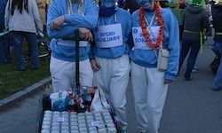 Drei Personen in blauen und weißen Kostümen, wahrscheinlich für eine Wohltätigkeitsveranstaltung, stehen mit einem Wagen voller Wasserflaschen. Dahinter gehen mehrere Personen auf einem Grasbereich.