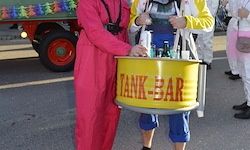 Zwei Personen in Kostümen halten einen gelben Eimer mit der Aufschrift 'Tank-Bar' in einem Außenbereich. Ein roter LKW ist im Hintergrund zu sehen.
