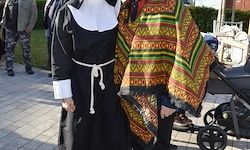 Eine Nonne in einem schwarzen Habit steht neben einer Person, die einen bunten Poncho trägt. Dahinter ist eine verschwommene Ansicht eines Kinderwagens und von Menschen, die auf dem Bürgersteig gehen.