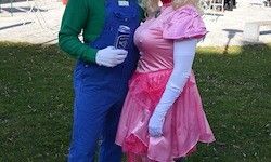 Ein Mann und eine Frau in Kostümen auf einem Grasfeld, wobei der Mann eine Dose hält und beide für die Kamera lächeln.