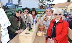 Eine Gruppe von Frauen in Kostümen und Sonnenbrillen steht hinter einem Tisch mit Körben und Flaschen darauf und einem Banner im Hintergrund.