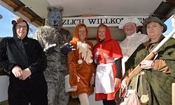 Eine Gruppe von Menschen in Kostümen posiert für ein Foto. Ein Wolfskostüm, ein roter Umhang und eine weiße Schürze sind zu sehen. Ein Schild mit der Aufschrift 'Zlich Willko' ist vorhanden.