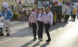 Drei Frauen in passenden rosa Oberteilen und schwarzen Hosen gehen die Straße entlang, mit einem Paradefahrzeug und Zuschauern im Hintergrund.