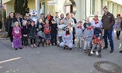 Eine Gruppe von Menschen, darunter Erwachsene und Kinder, in verschiedenen Kostümen gekleidet, stehen und hockend auf der Straße vor einem Gebäude. Einige tragen Brillen und Mützen.