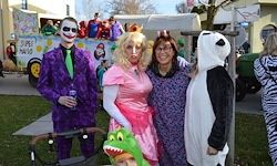Eine Gruppe von Menschen in verschiedenen Kostümen posiert für ein Foto auf einem Rasenfeld. Ein Mann in einem Joker-Kostüm, eine Frau in einem rosa Kleid und eine weitere Frau in einem Panda-Kostüm stehen in der Nähe eines Kinderwagens.