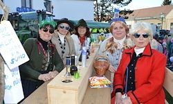 Fünf Frauen stehen an einem Holztresen, geschmückt mit Partyhüten und Sonnenbrillen. Flaschen und eine Schüssel sind auf dem Tresen, und ein Schild hängt im Hintergrund.