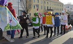 Eine Gruppe von Menschen in bunten Kostümen und Hüten, die während einer Parade auf der Straße gehen. Im Hintergrund sind Gebäude und ein Baum zu sehen.
