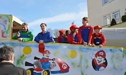 Eine Gruppe von Kindern in Mario-Kostümen steht auf einem dekorierten Wagen. Ein Mann steht auf der linken Seite. Ein Gebäude ist im Hintergrund.