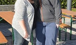Zwei Personen stehen zusammen in einem Biergarten mit Holzbänken, die Frau in einem grauen Cardigan und der Mann in einem grauen Hoodie.