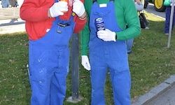 Zwei Personen in blauen Overalls und weißen Handschuhen stehen in einem Grasbereich. Einer trägt ein rotes Hemd und der andere ein grünes. Sie halten Flaschen und es gibt einen verschwommenen Hintergrund von Menschen und einem Fahrzeug.