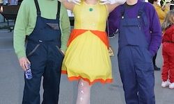 Drei Menschen in Kostümen posieren für ein Foto auf der Straße. Eine Person trägt ein gelb-oranges Kleid, während die anderen beiden blaue Latzhosen tragen. Ein Kind in Rot steht im Hintergrund.