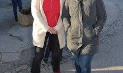 Zwei Frauen stehen auf einer rissigen Straße, eine in einem roten Shirt und einer weißen Strickjacke, die andere in einer grauen Jacke und Jeans. Hinter ihnen ist teilweise eine Person in blauen Jeans und Turnschuhen zu sehen.