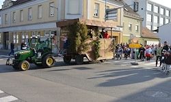 Ein grüner Traktor, mit Zweigen dekoriert, fährt die Straße hinunter, mit Menschen und Gebäuden im Hintergrund.