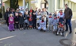 Eine Gruppe von Menschen in Kostümen, einige mit Rentier-Kopfbändern, posiert für ein Foto vor einem Gebäude. Ein Mann in einem schwarzen Mantel steht rechts.