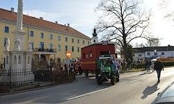 Ein grüner Traktor zieht einen roten Wagen auf einer Straße mit Gebäuden und Bäumen. Menschen gehen vorbei und stehen herum.