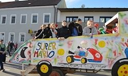 Ein geschmückter Wagen mit den Worten 'Super Mario' darauf, gefüllt mit Menschen vor einem Gebäude mit vielen Fenstern.