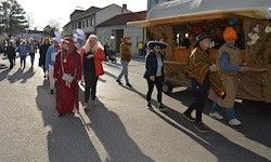 Eine Straße mit Menschen in Kostümen, einer mit einem Turban, geht im Schatten eines farbenfrohen Wagens. Im Hintergrund Gebäude und Bäume.