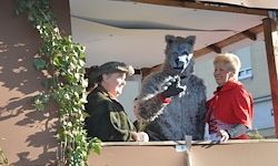 Drei Personen stehen auf einem Balkon, zwei in Winterkleidung, eine mit einem Wolfskostüm, während sie lachen und mit dem Publikum interagieren.