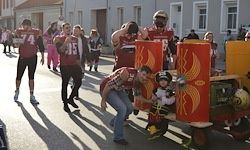 Eine Gruppe von Menschen in Footballtrikots und Helmen zieht einen Karren mit einem Kind auf der Straße. Dahinter gehen weitere Menschen in die gleiche Richtung, einige in rosa Kostümen. Gebäude säumen die Straße.