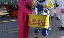 Eine Person in einem rosa Kostüm trägt einen gelben Fass, der mit 'TANK-BAR' beschriftet ist. Eine weitere Person in einem gelb-blauen Outfit hält eine Tasche. Dahinter ist ein Fahrzeug mit einem roten Rad zu sehen.