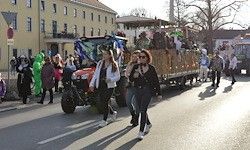 Menschen gehen an einem von einem Traktor gezogenen Umzugswagen vorbei. Mehrere Personen sitzen auf dem Wagen. Andere gehen am Straßenrand entlang und tragen Kostüme.