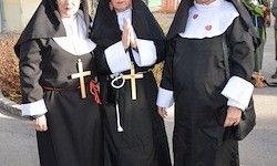 Drei Nonnen stehen zusammen, gekleidet in schwarze Kutten mit weißen Akzenten. Zwei von ihnen haben hölzerne Kreuze auf der Brust.