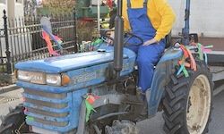 Eine Person in gelbem Hemd und blauer Latzhose sitzt auf einem blauen Traktor, der auf der Straße geparkt ist. Es sind bunte Bänder am Traktor befestigt.