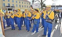 Eine Musikkapelle in gelben und blauen Kostümen spielt Blasinstrumente im Freien. Im Hintergrund befinden sich Gebäude und Bäume.