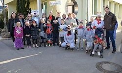Eine Gruppe von Menschen, in verschiedenen Kostümen gekleidet, posiert für ein Foto vor einem Gebäude. Einige tragen Winterkleidung, andere haben Gesichtsbemalung. Ein Mann steht in der Nähe eines Kinderwagens.