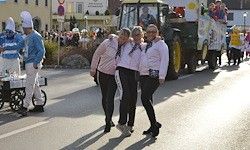 Drei Frauen stehen auf der Straße, ein Traktor im Hintergrund. Ein Umzug findet auf der Straße statt, bei dem die Menschen Kostüme tragen.