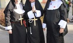 Drei Nonnen in schwarzen Gewändern mit weißen Ärmeln und Kreuzen auf der Brust stehen zusammen auf einer Straße.