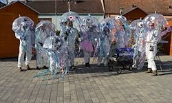 Eine Gruppe von Menschen in weißen Kostümen mit Regenschirmen auf einem gepflasterten Boden vor Häusern.