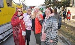 Vier Frauen in Winterkleidung stehen auf einer Straße, zwei tragen rote Mäntel, eine trägt einen rosa Mantel und eine andere einen silbernen Puffermantel. Ein gelber Bus steht hinter ihnen.